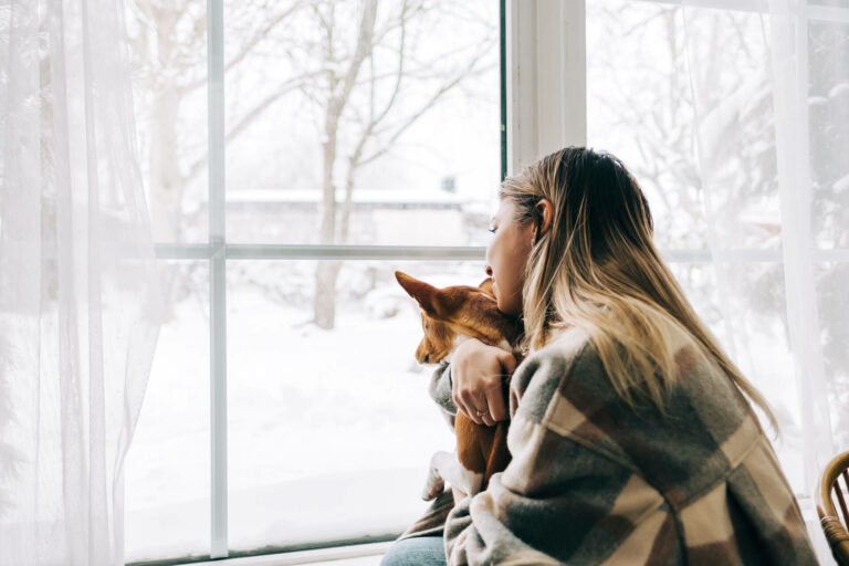 A woman and her dog in front of the windows looking at the snow outside.